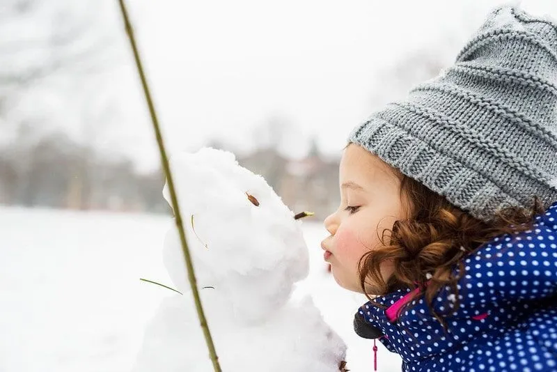 Little girl wearing a grey woolly hat, standing outside kissing a snowman.