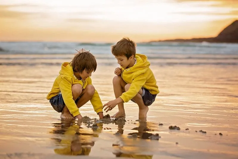 Two young boys wearing matching yellow hoodies, crouching down to play with the sand on the beach.