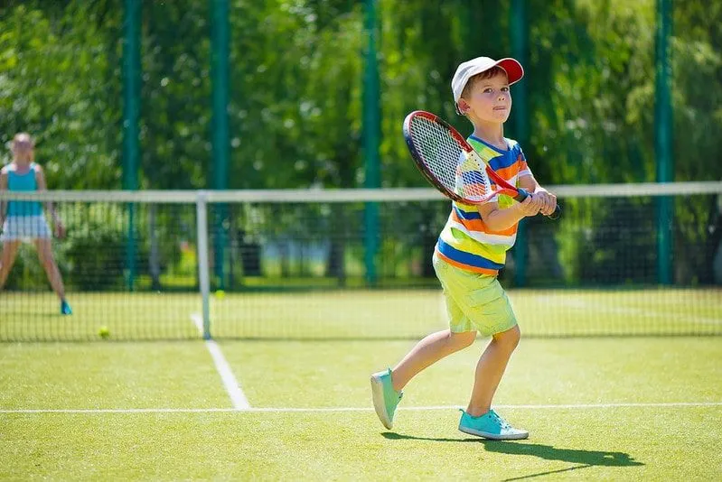 Young boy in a colourful striped t shirt and cap playing tennis.