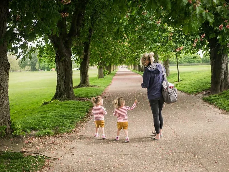 Little twins walking in the park with their mum under a tree-lined path on a day out in Chelmsford.