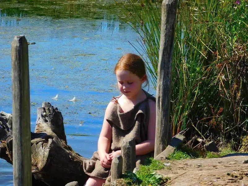 Viking girl sat at the edge of the water of a pond, looking down at her hands.