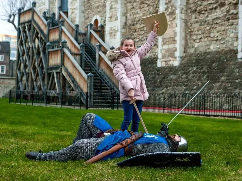 Girl playfully acting out defeating a man in armour and chain mail, as he lies on the ground her wooden sword at his chest.