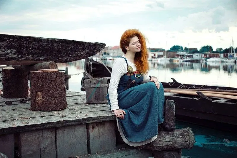 Woman sat by a Viking boat next to the water, wearing Viking clothing.