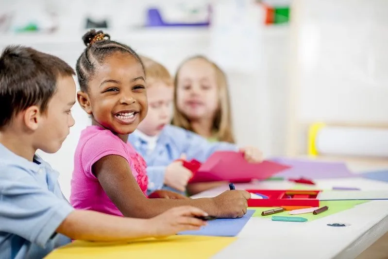 Kids sat at a table smiling, colouring on pieces of coloured paper.