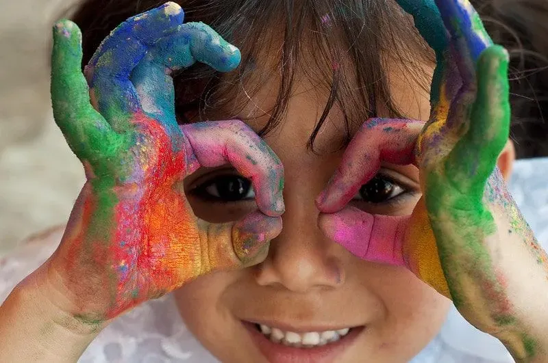 Little girl with colourful paint on her hands, making a face and smiling.