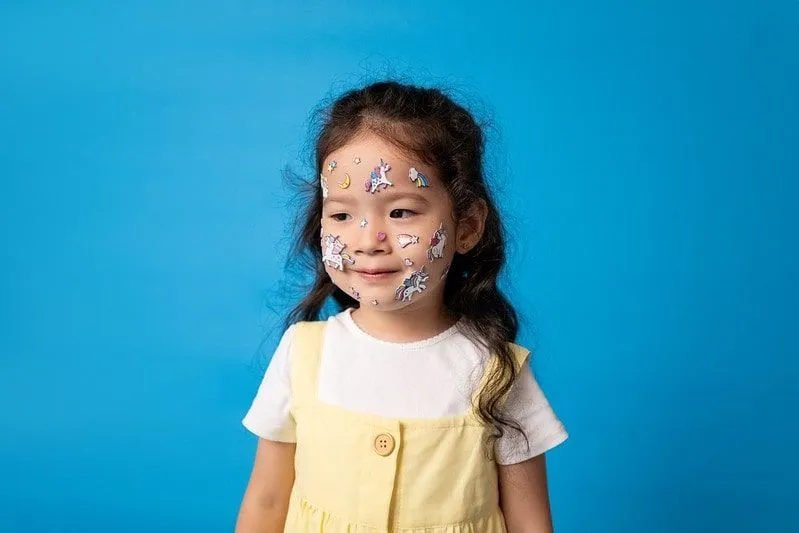 Little girl with unicorn stickers stuck on her face, standing in front of a blue background.