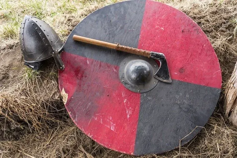 Red and black wooden Viking shield, with an axe and helmet resting against it.