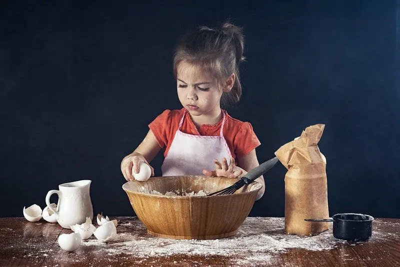 Little girl wearing a pink apron baking a cake, breaking an egg into the bowl.
