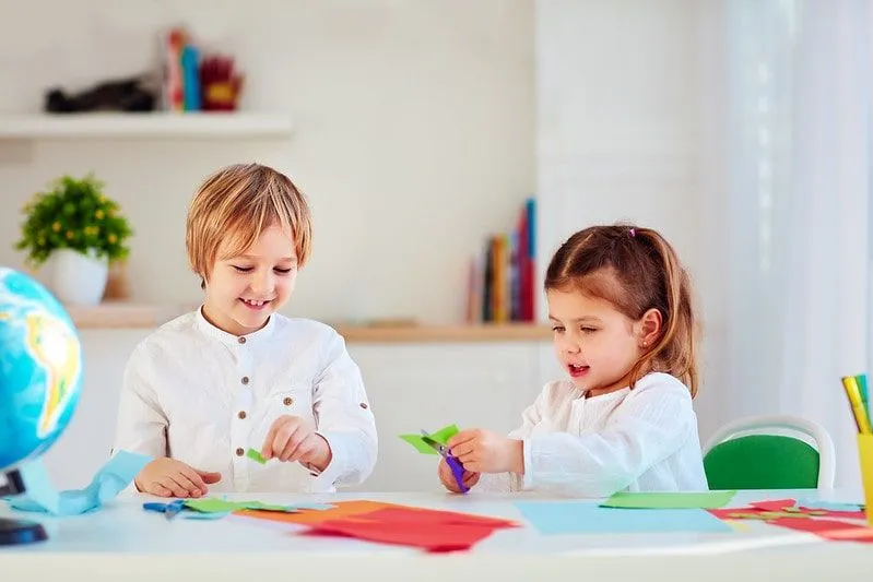 Two kids sat at the table cutting paper to make an origami 