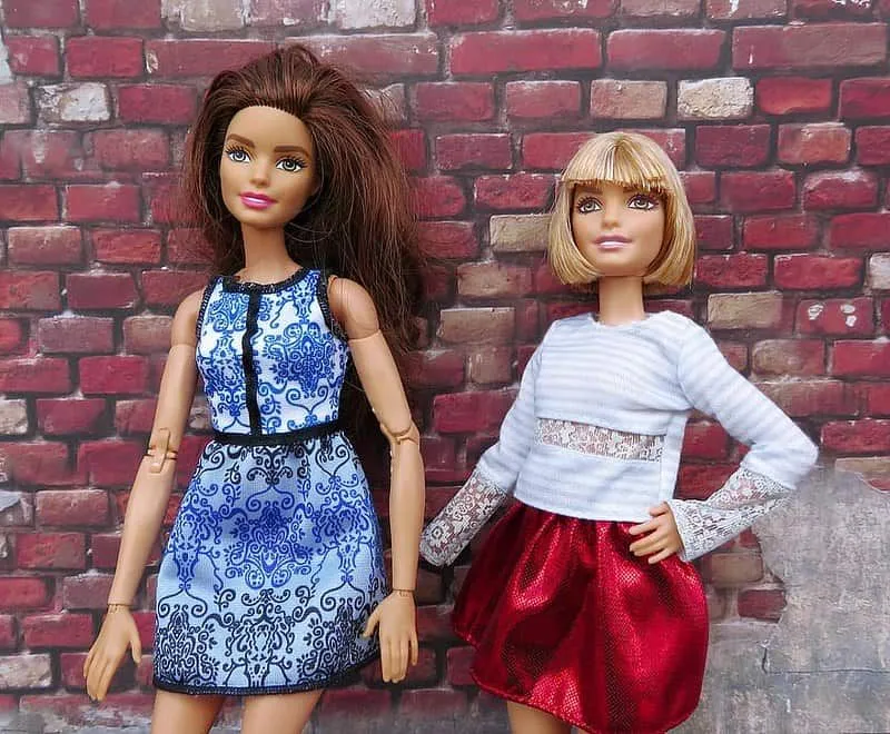 Two Barbie dolls striking a pose in front of a red brick wall.