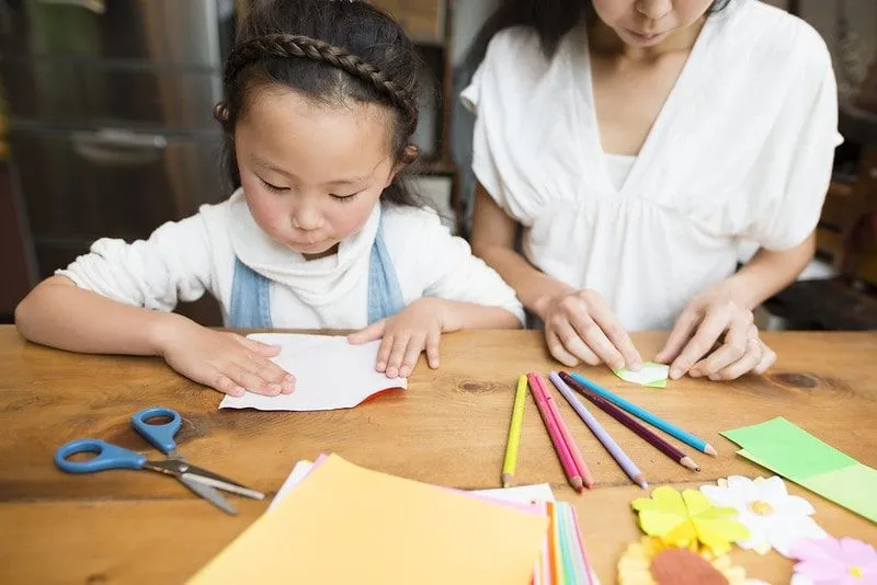 Young girl and her mum at the table cutting out paper to make dolls shoes.