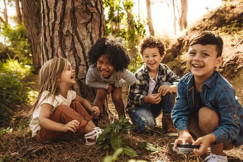 Young kids sitting in the forest next to a tree laughing at plant jokes.