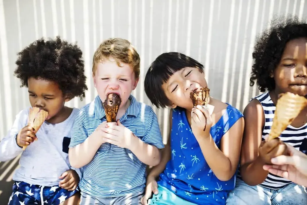 Four kids stood together licking their ice cream cones with ice cream all around their mouths.