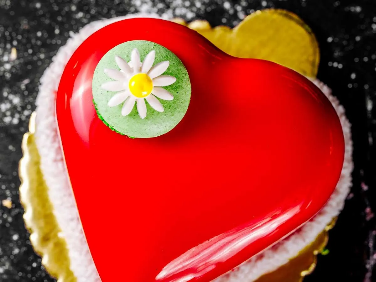 Red heart shaped cake with a green macaron with a daisy on top, on top of the cake.