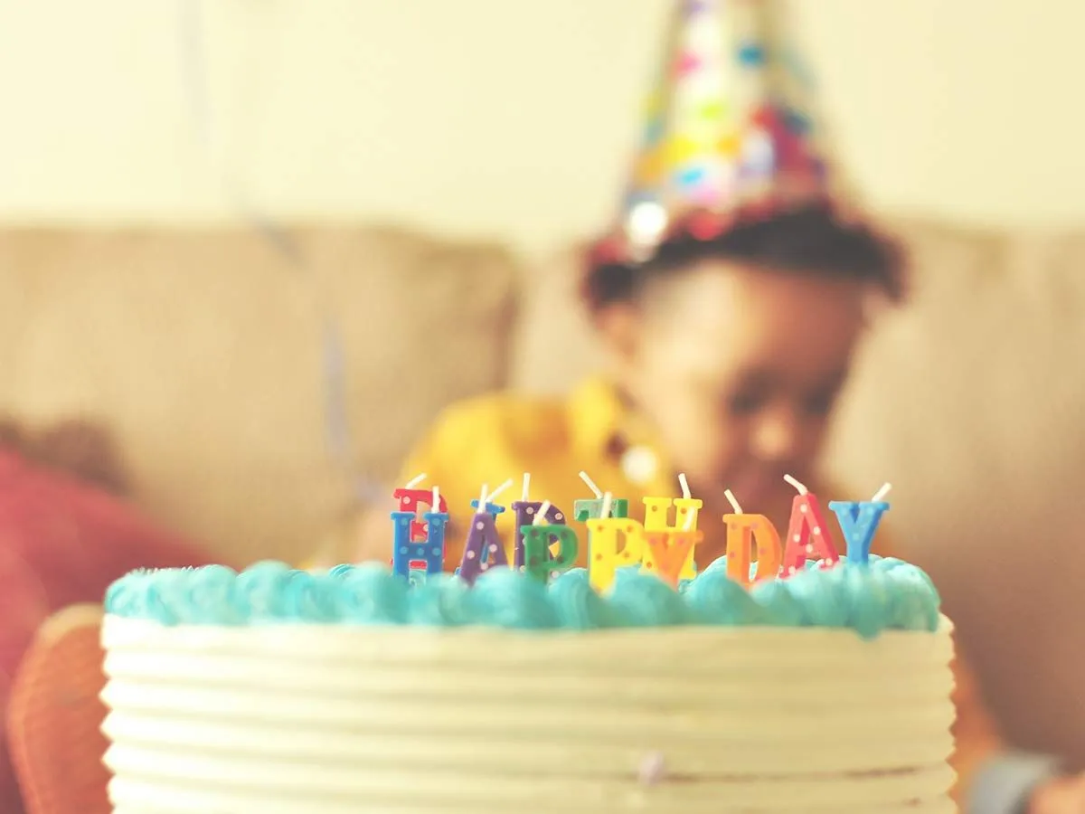 Birthday cake with candles on the table, a toddler wearing a party hat sat behind in the background.