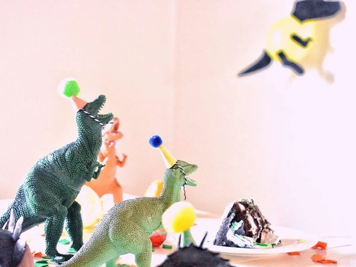 Toy dinosaurs wearing party hats on a table surrounding a piece of birthday cake.