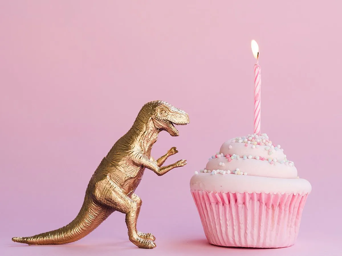 Gold dinosaur toy facing a pink cupcake with a lit candle in it.
