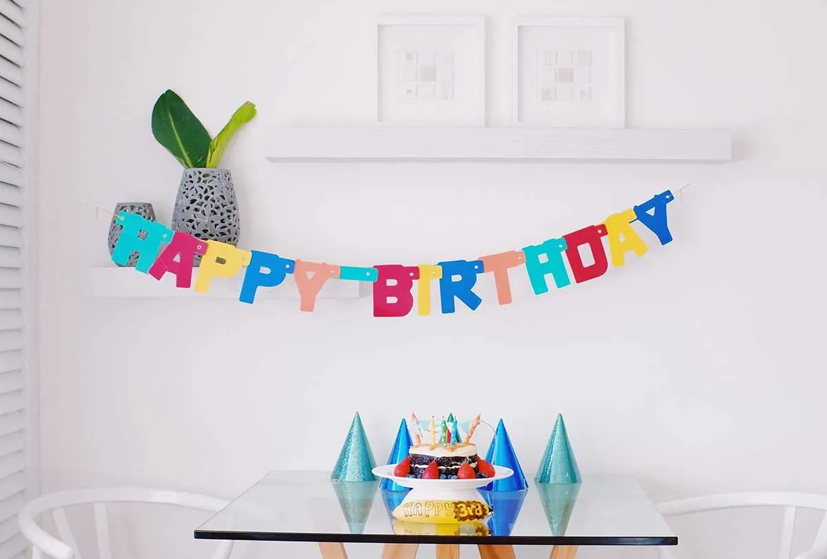 Happy birthday spelled out in a banner hanging on the wall behind a table with a cake and party hats on it.