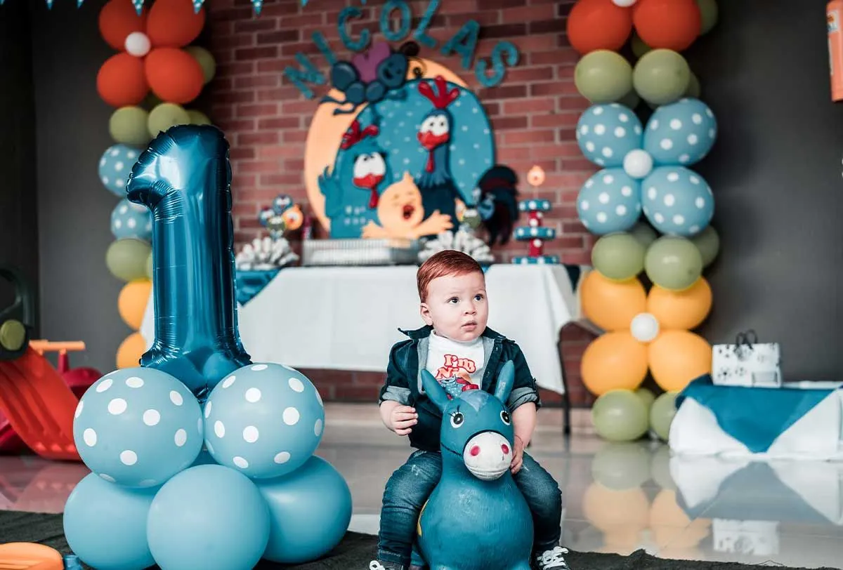 Toddler sat on a toy donkey at his first birthday party with balloon banners decorating.
