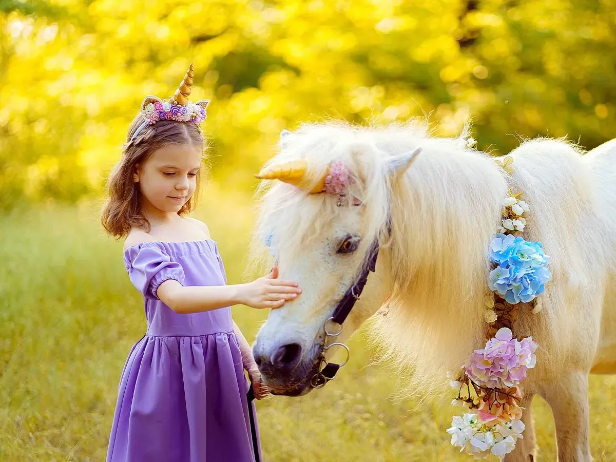 Young girl in a purple dress and DIY unicorn horn petting a white pony who is also wearing a unicorn horn.