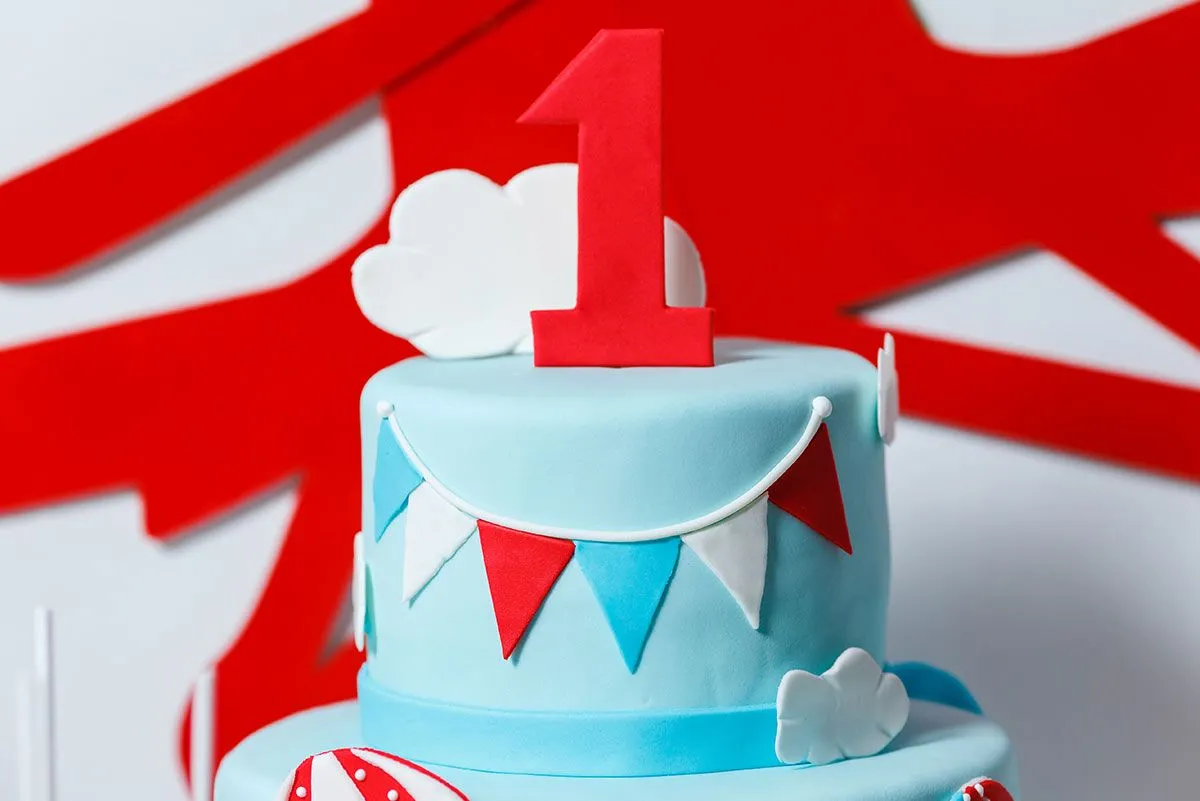 Blue, tiered first birthday cake with a big, red airplane decoration on the wall behind.