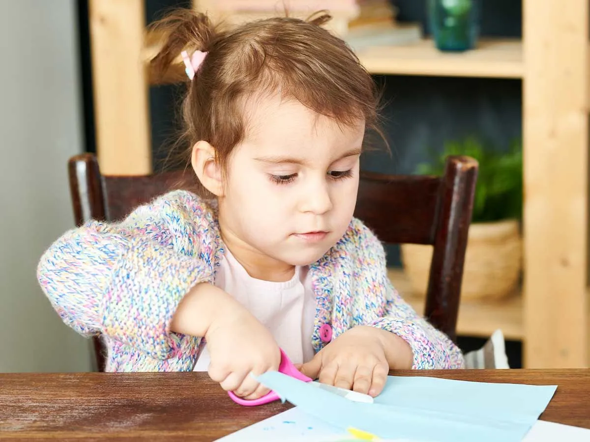 Little girl at the table cutting a piece of blue paper to make an origami eagle.