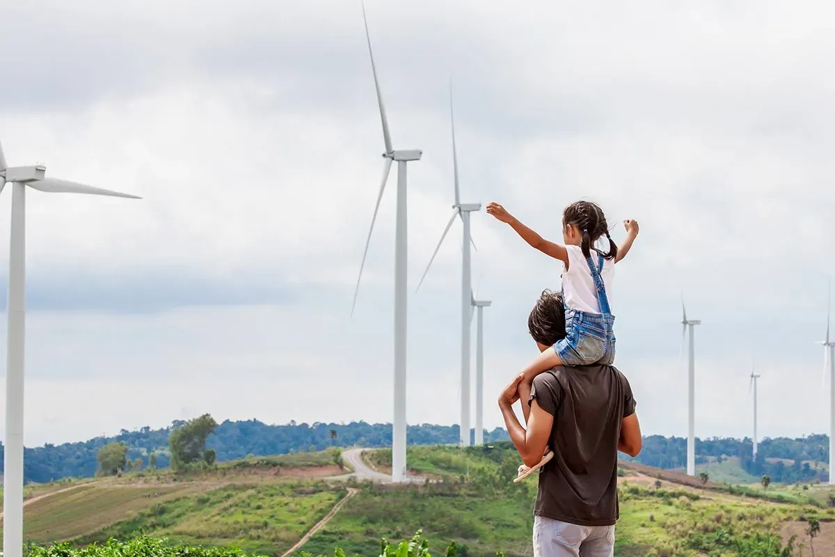 Little girl on her dad's shoulders standing on a hill looking at the wind turbines.