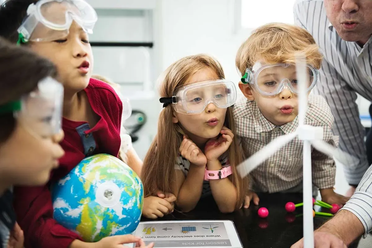 Young children in science class wearing goggles looking at a model of a wind turbine.