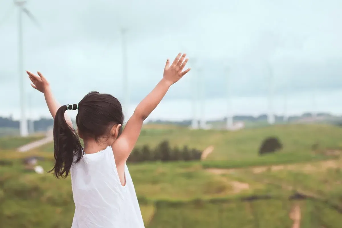 Little girl standing on a hill with her hands in the air, looking at the wind turbines.