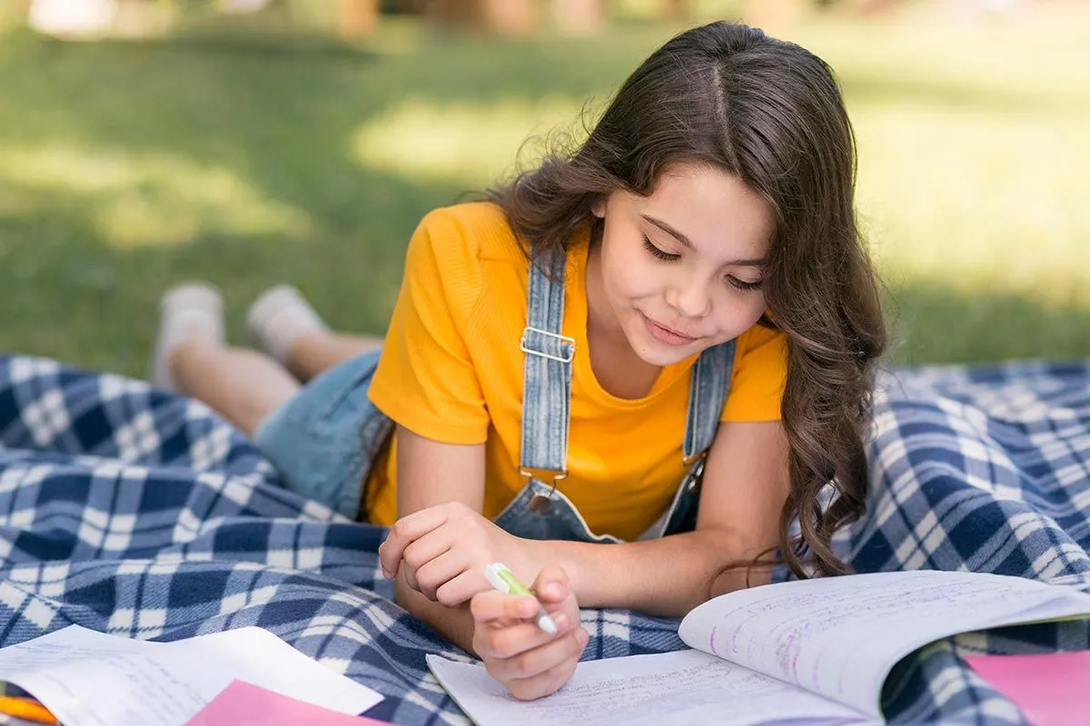 Young girl lying on her stomach on a rug outside, learning about hyphens writing in her workbook.