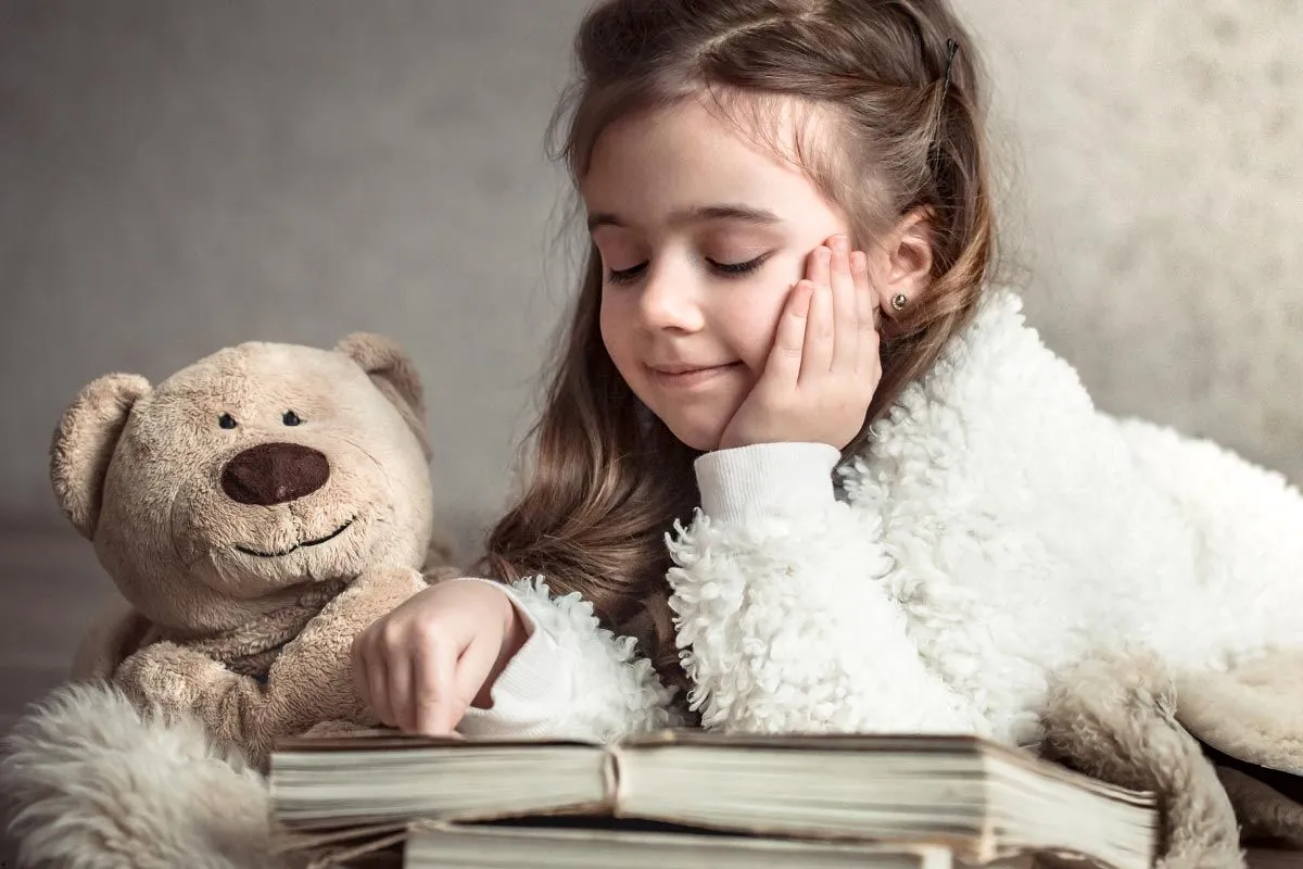 Young girl reading a book next to her brown teddy bear.