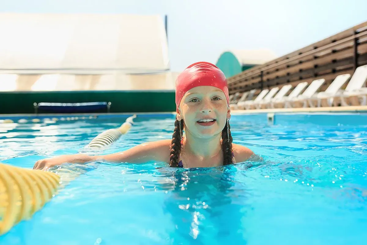 Young girl, wearing a red swimming cap, swimming in an outdoor pool.