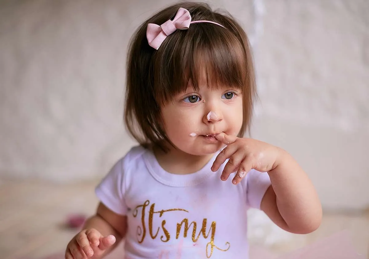 Baby girl licking cake off her finger, with more icing on her nose.
