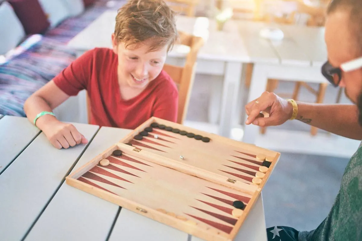 Teenage boy sat at a table outside playing backgammon with his dad.
