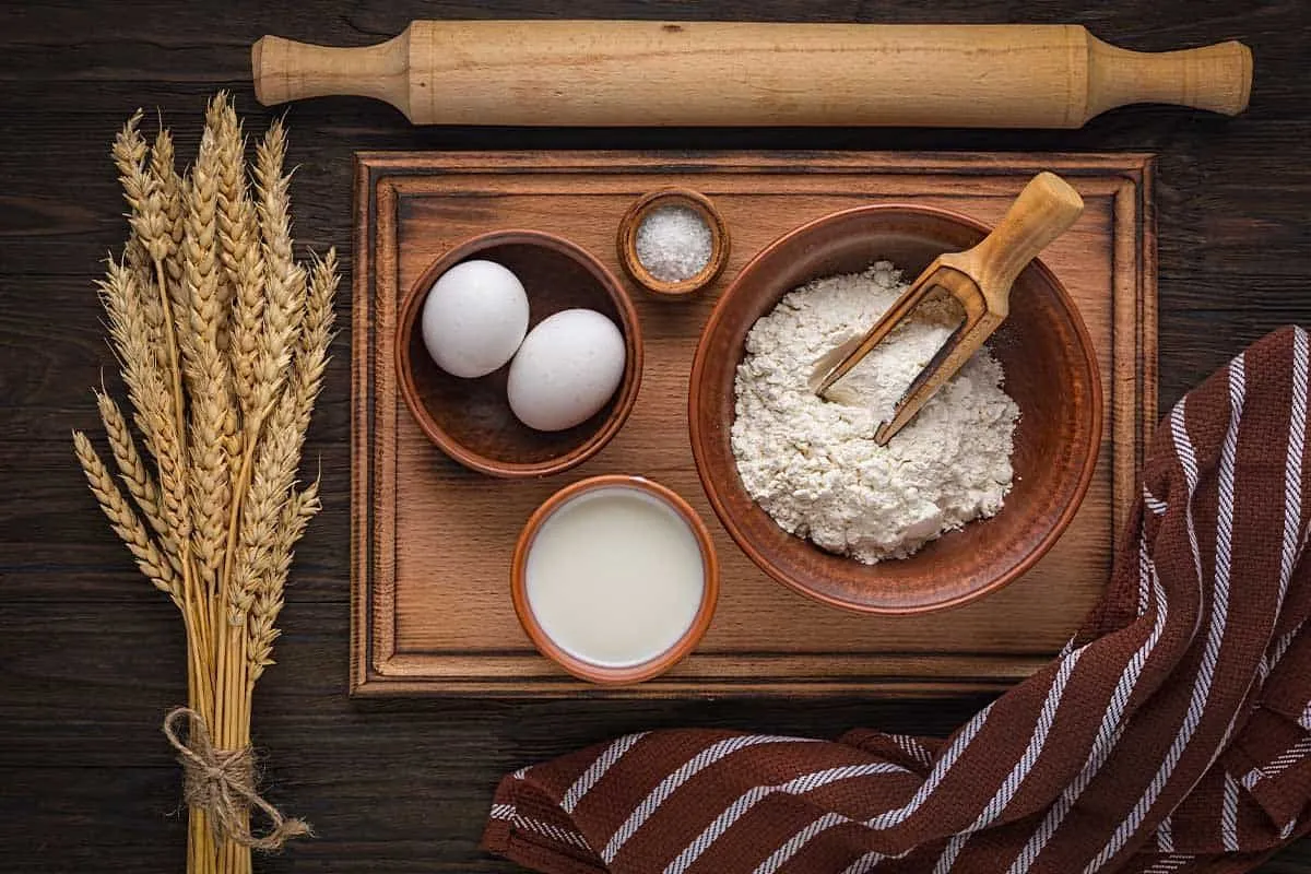 A rolling pin and a chopping board with flour, milk and eggs laid out on it, ready to make a pizza cake.