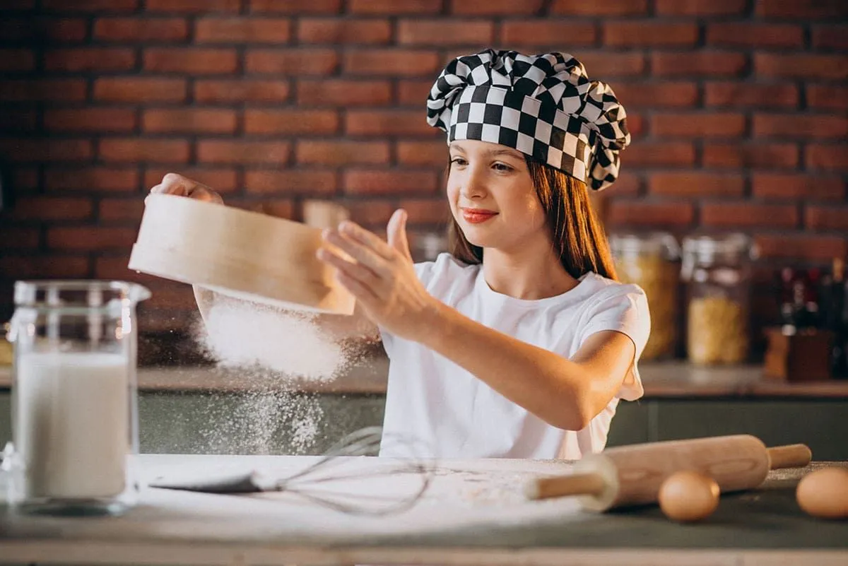 A young girl sifting flour to make a pizza cake, she is wearing a black and white chef's hat.