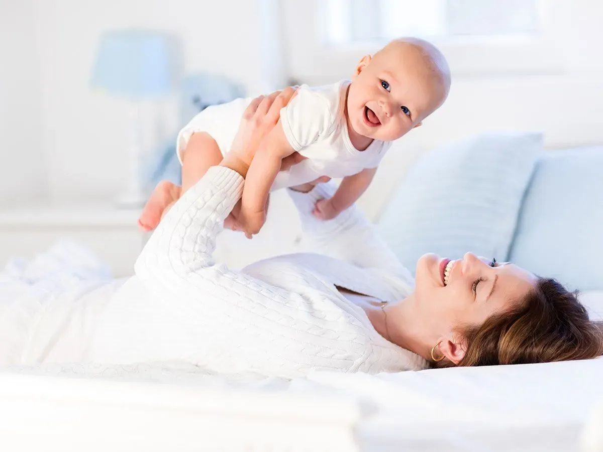 A mother is lying on the bed lifting her smiling baby above her into the air.