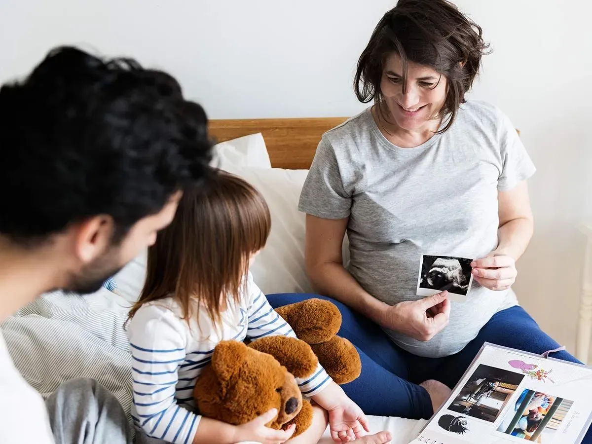 A family are looking through a baby scrapbook together, the pregnant mother smiles as she shows a baby scan photo to the rest of the family.