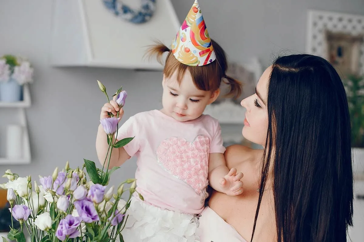 A mum holds and looks at her toddler who is playing with some flowers whilst wearing a birthday party hat.