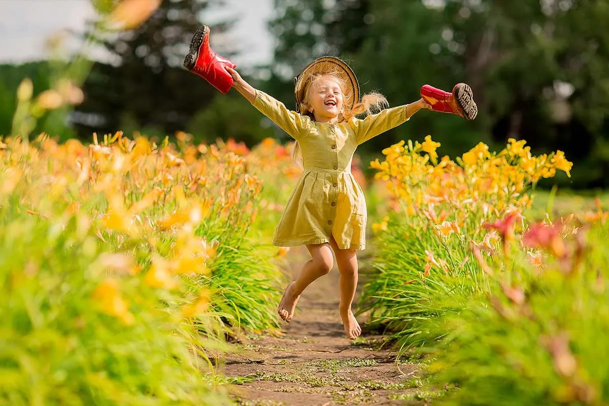 A happy young girl in a yellow dress runs through a field of yellow flowers holding wellies in her hands.