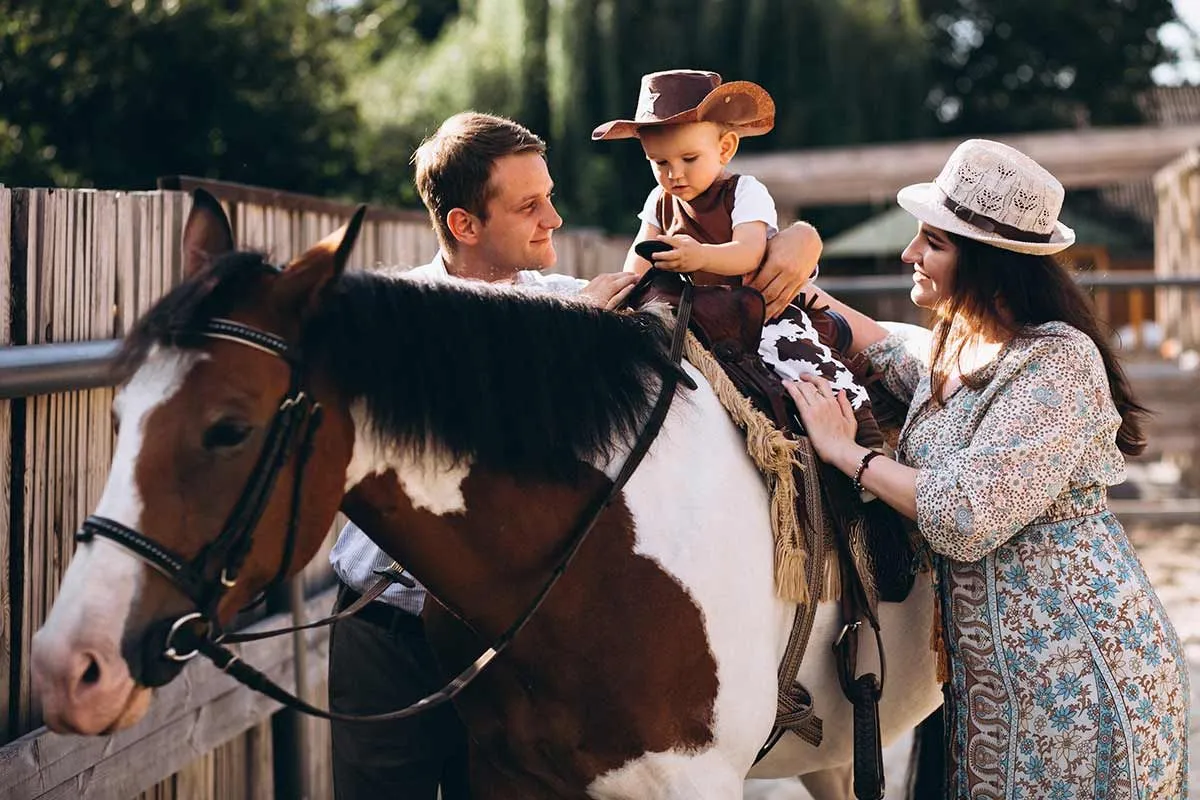 A young boy wearing a cowboy hat rides a brown and white horse whilst his parents support him from either side.