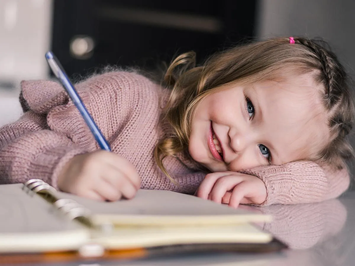 A little girl writing in a notebook looks up and smiles at the camera.