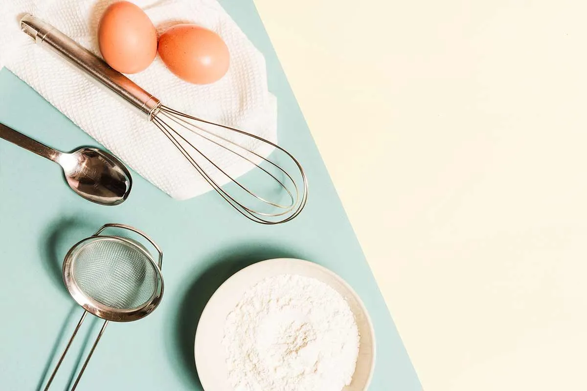 Two eggs, a bowl of flour, a sieve, a whisk and a spoon laid out on a blue and white table.