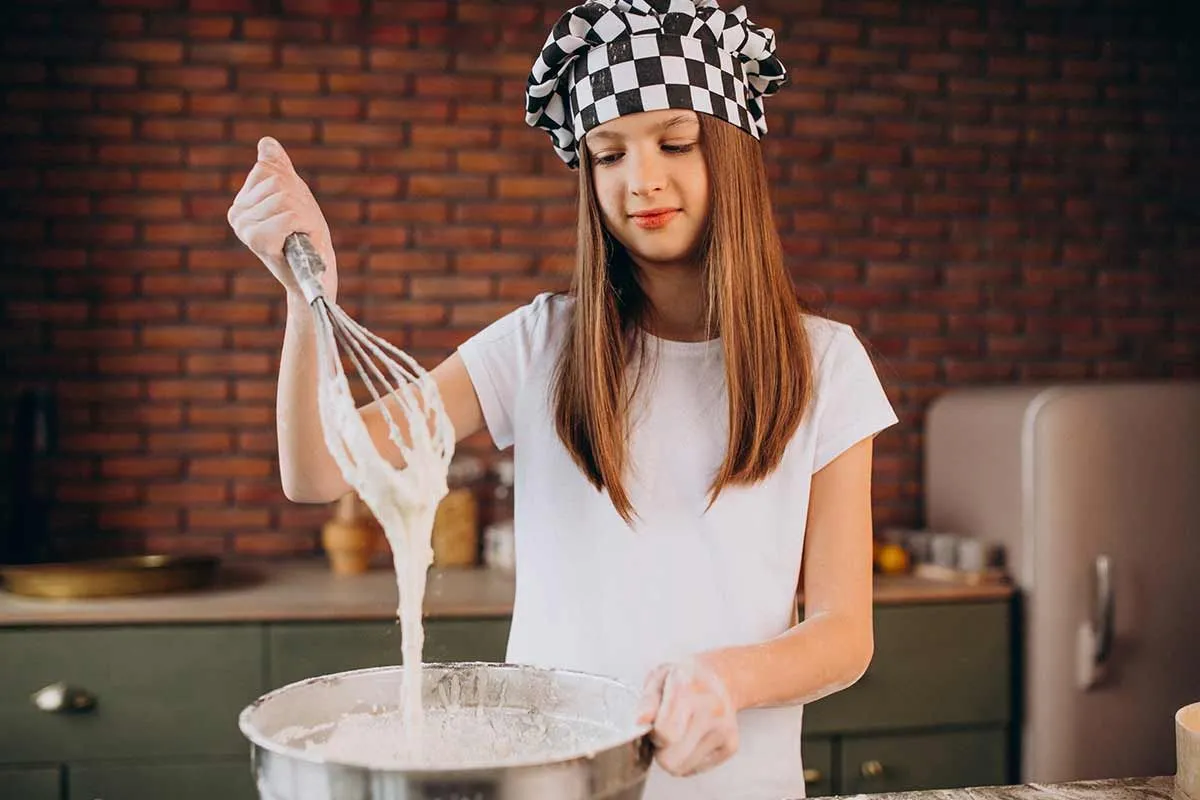 A teenage girl wearing a chef's hat whisks together ingredients in a bowl to make a leopard cake.