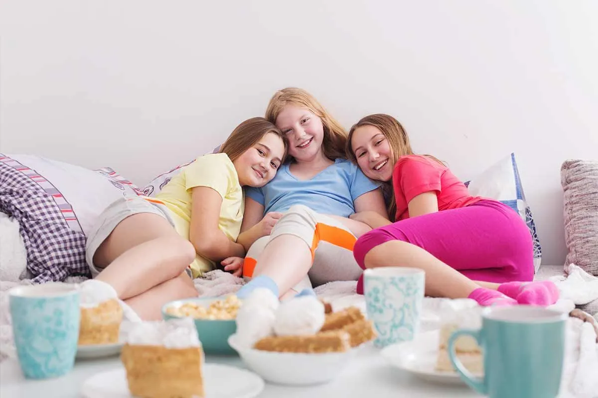 Three teenage girls sit on a sofa smiling, in front of them is a table with tea and cake laid out.