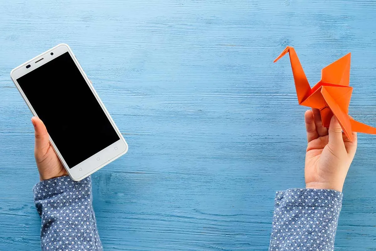Close up of a child's hands holding an orange origami bird in one and a smartphone in the other.