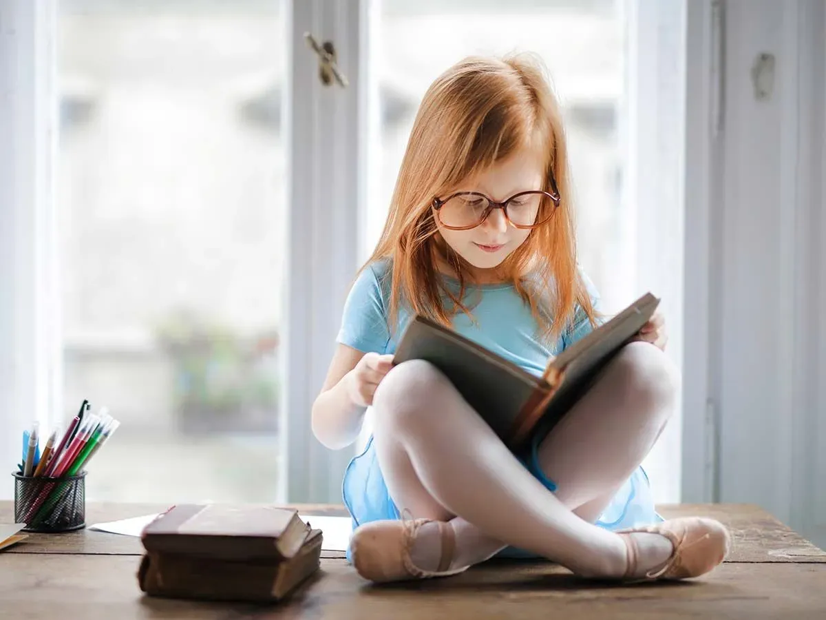 Little girl wearing glasses sat cross-legged on the table reading a book about synonyms and antonyms.