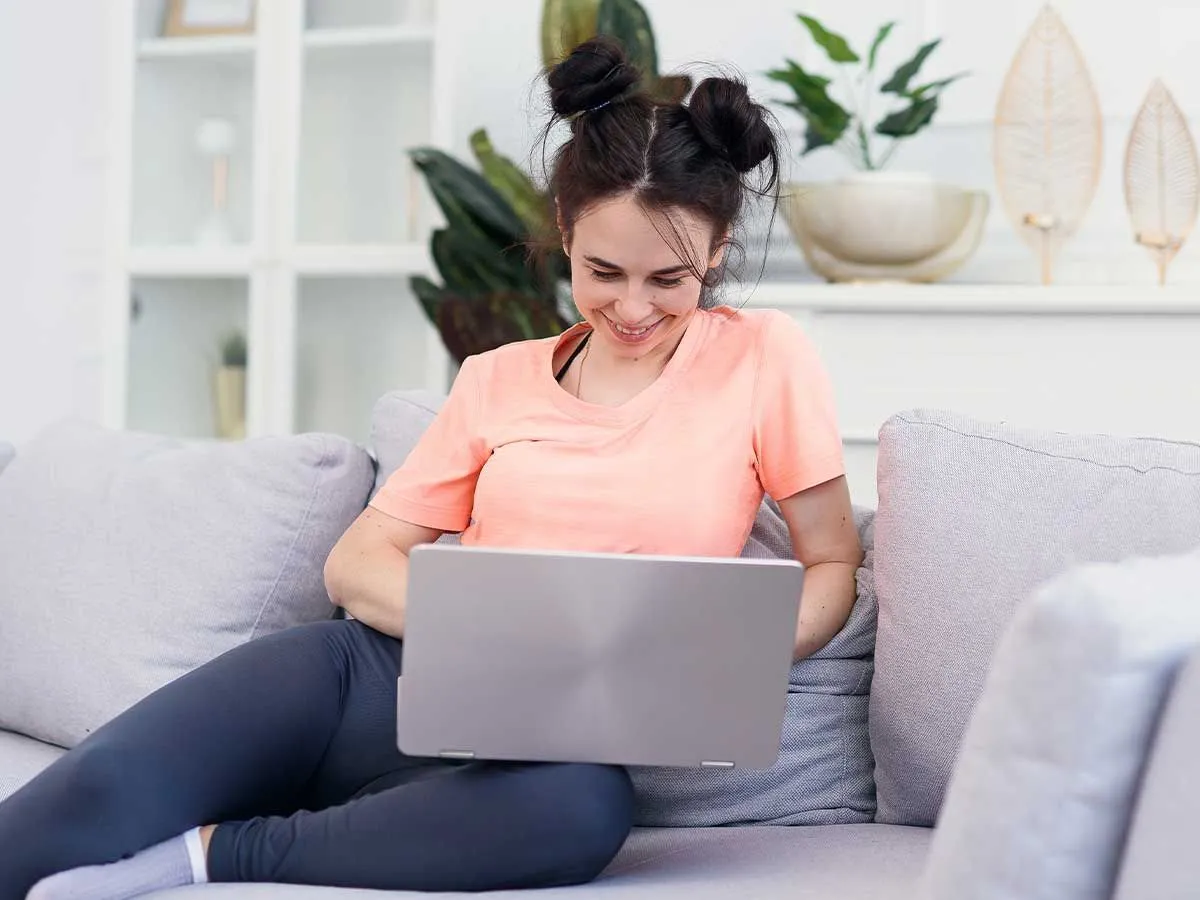 A teenage girl in an orange t-shirt sits on the sofa using her laptop, she is smiling.