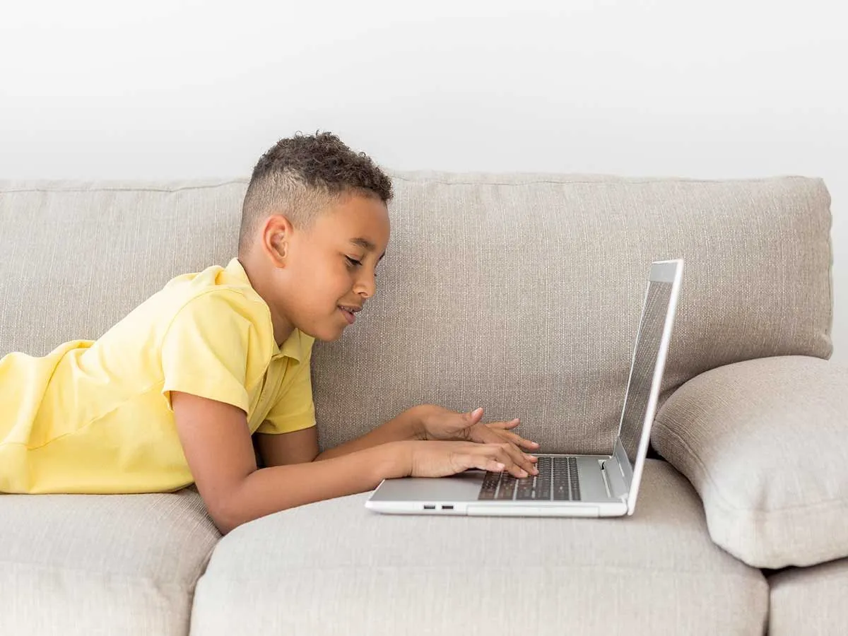 A tween boy lies on a grey sofa, he is smiling and typing on his laptop.