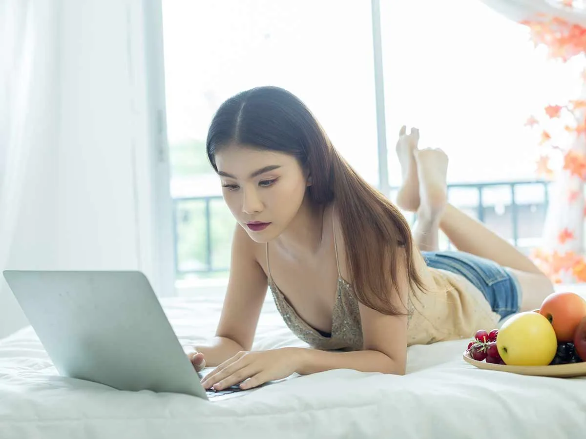 A teenage girl lies on her bed looking at her laptop, next to her is a fruit bowl.